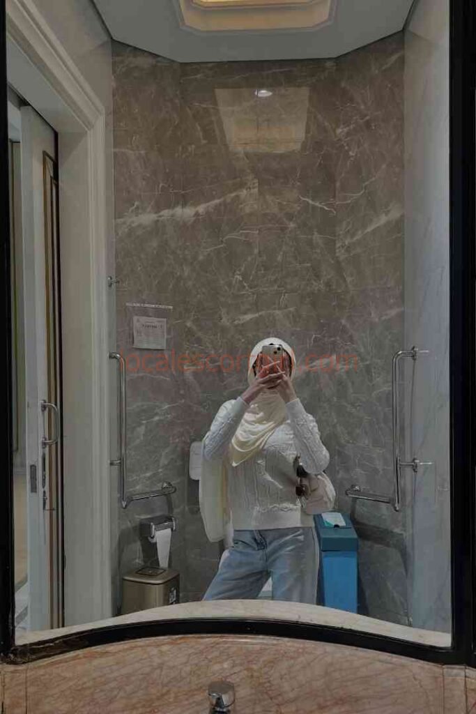 Young woman in hijab and white sweater taking mirror selfie in modern bathroom